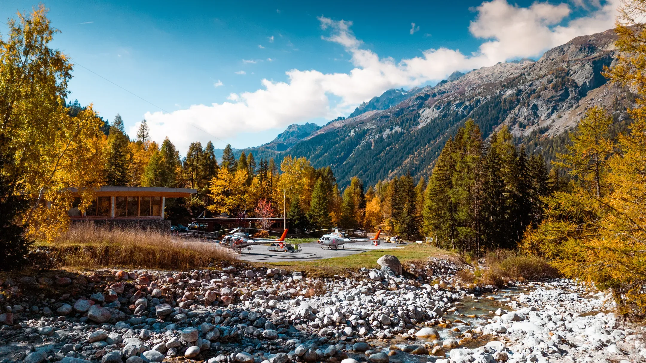 Base d'hélicoptères au bord d'une rivière, mélèzes jaunes et sommets