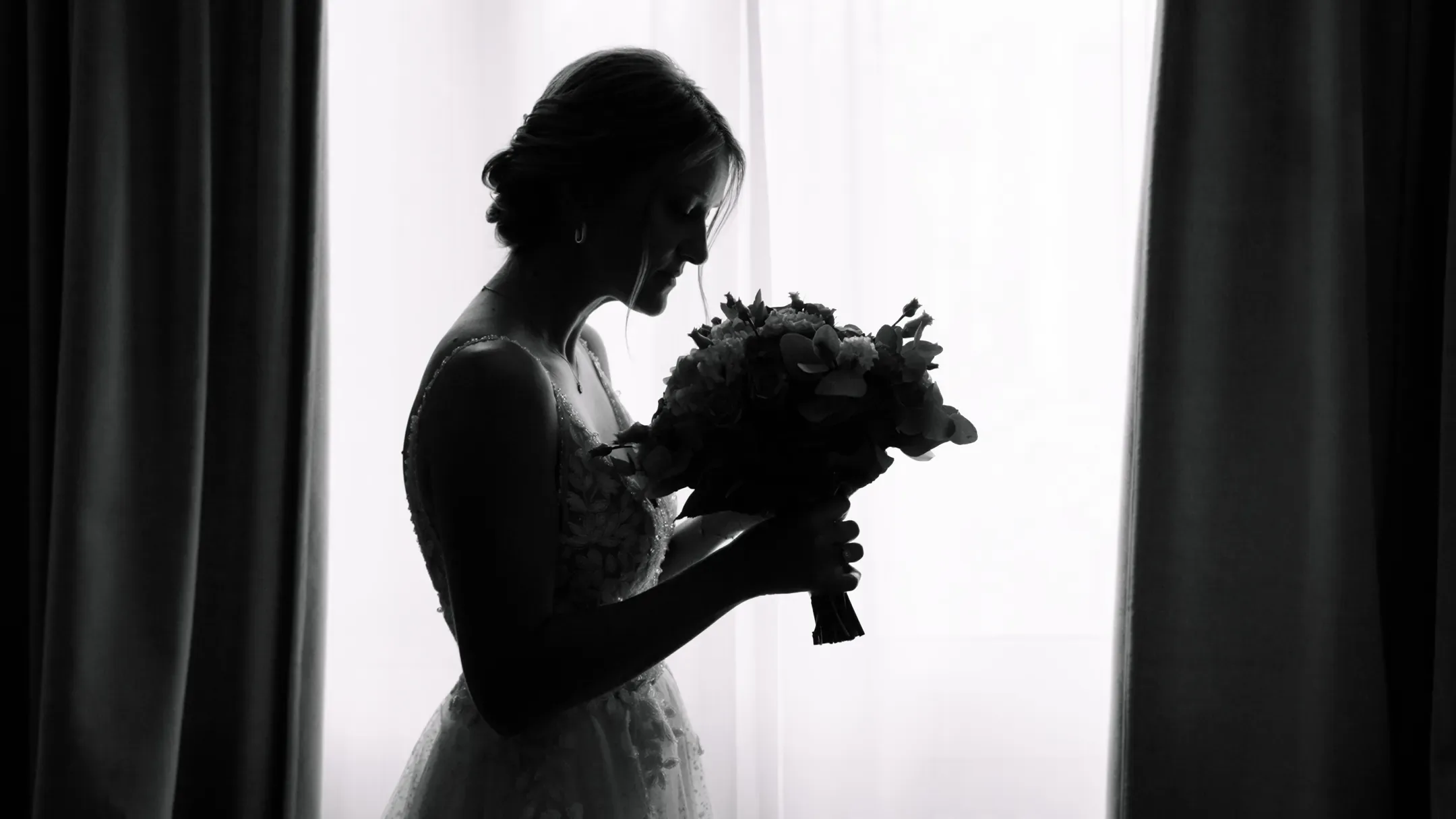 Silhouette de la mariée tenant son bouquet devant une fenêtre lumineuse, en noir et blanc