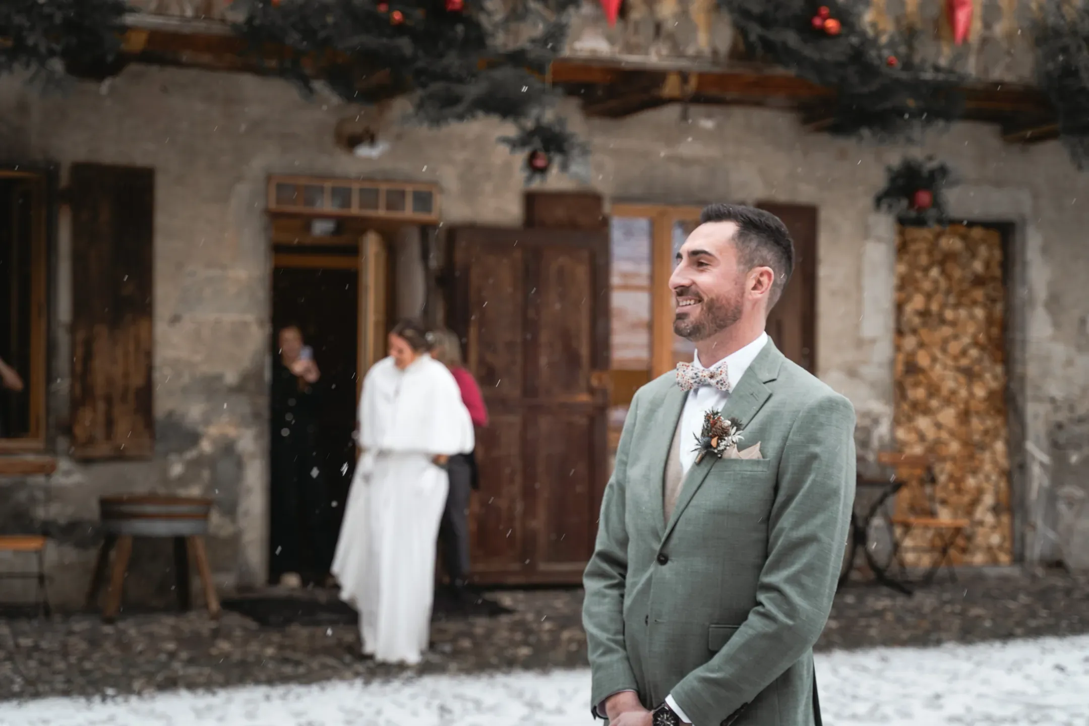 Le marié en costume vert devant un chalet enneigé, la mariée à l'arrière-plan