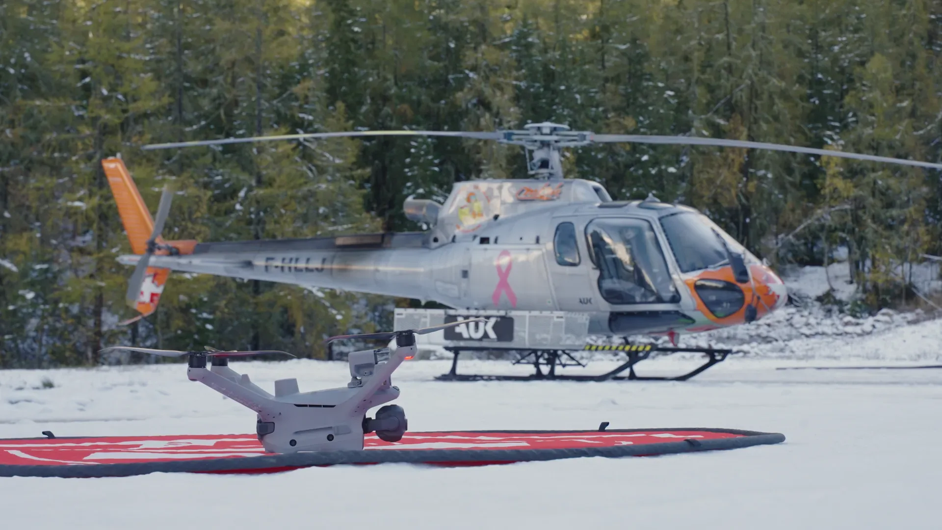 Drone sur sa plateforme dans la neige, hélicoptère posé à l'arrière-plan