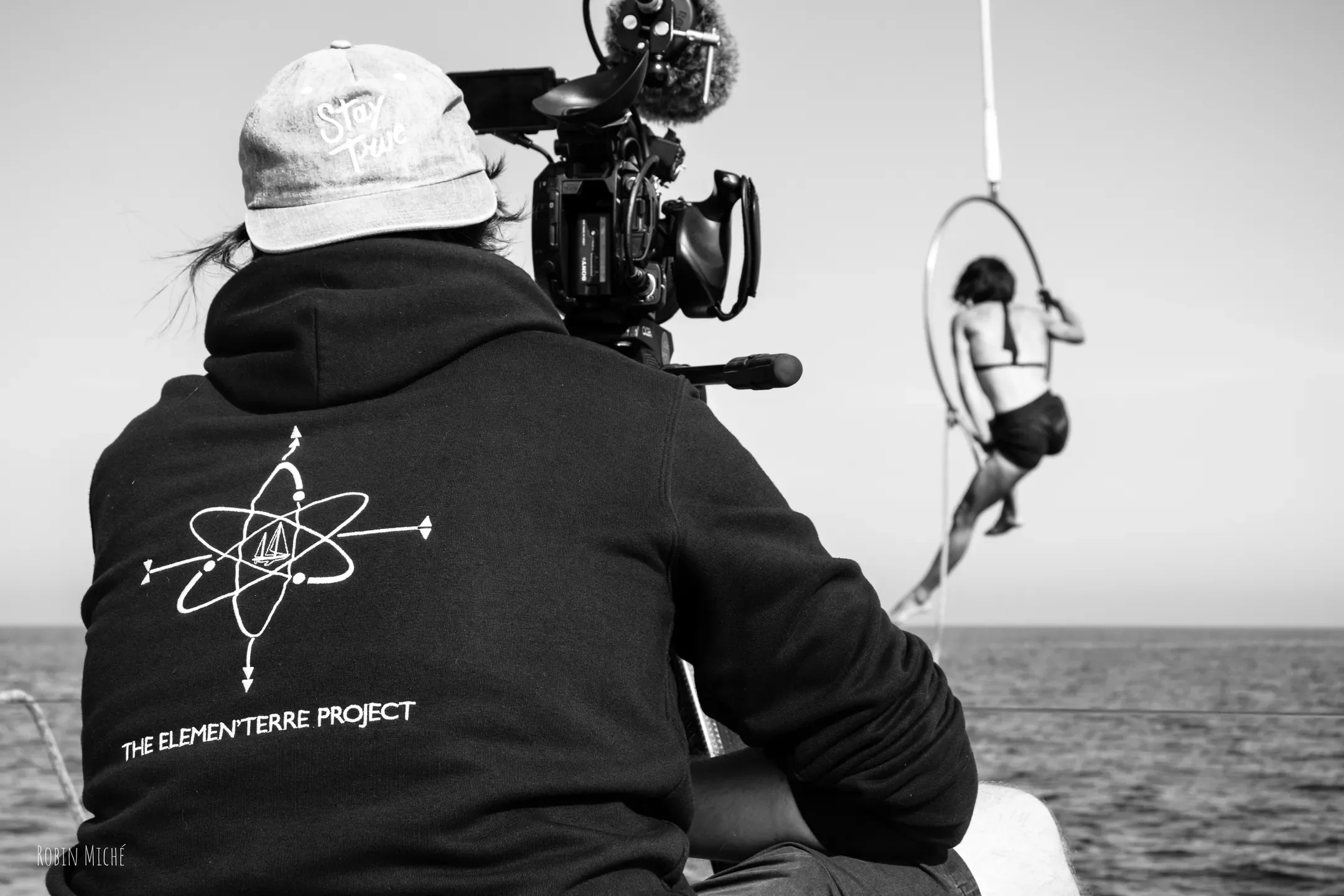 Cadreur de dos filmant le funambule suspendu au-dessus de la mer, en noir et blanc