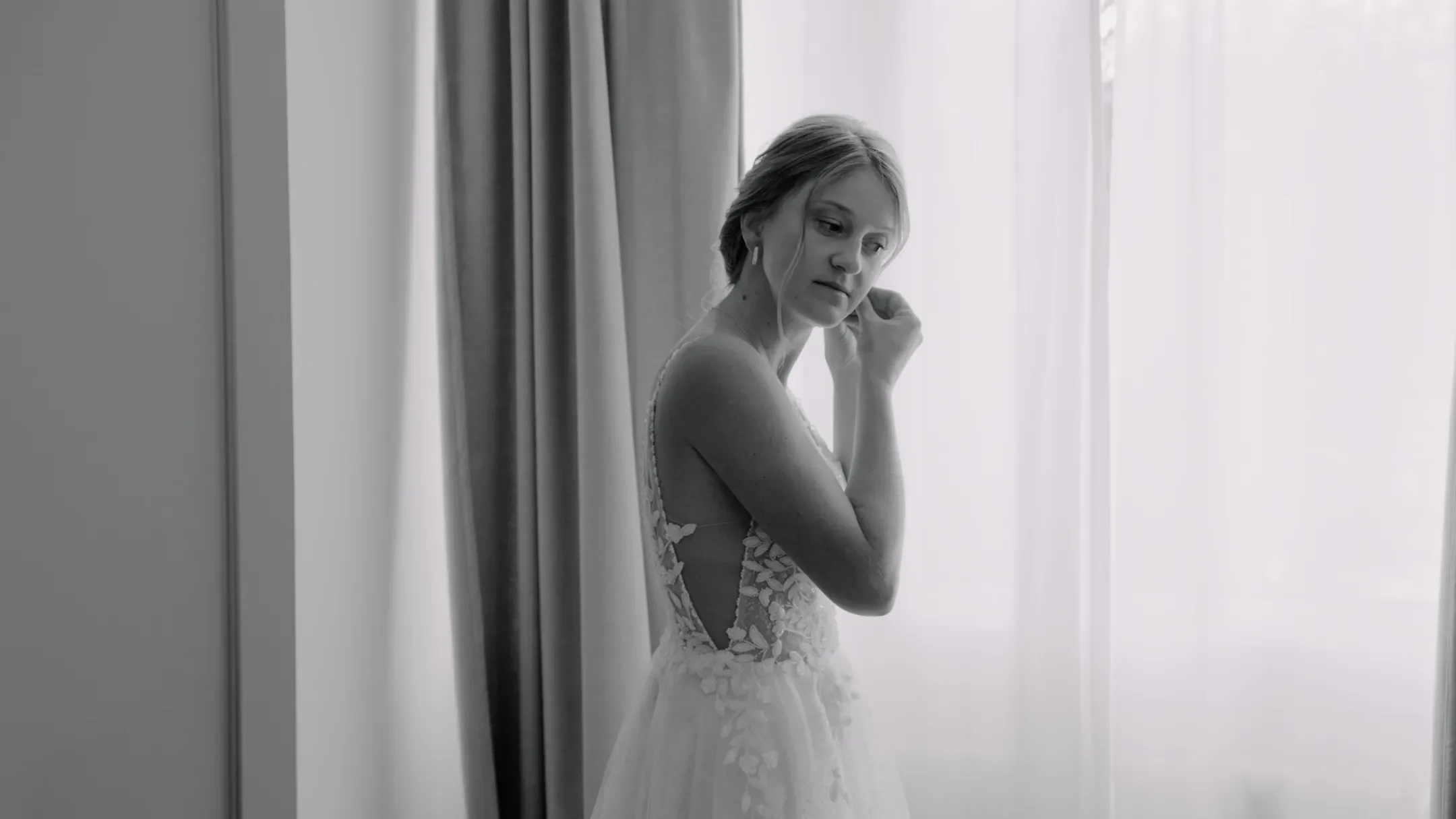 La mariée ajuste sa boucle d'oreille près d'une fenêtre, en noir et blanc