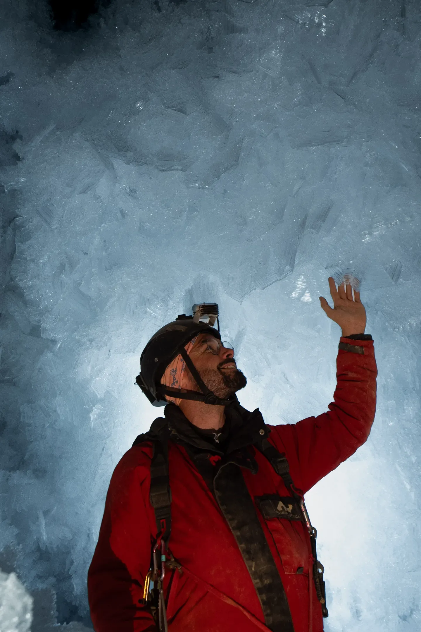 Spéléologue en combinaison rouge, la main posée sur une paroi de glace éclairée à la frontale