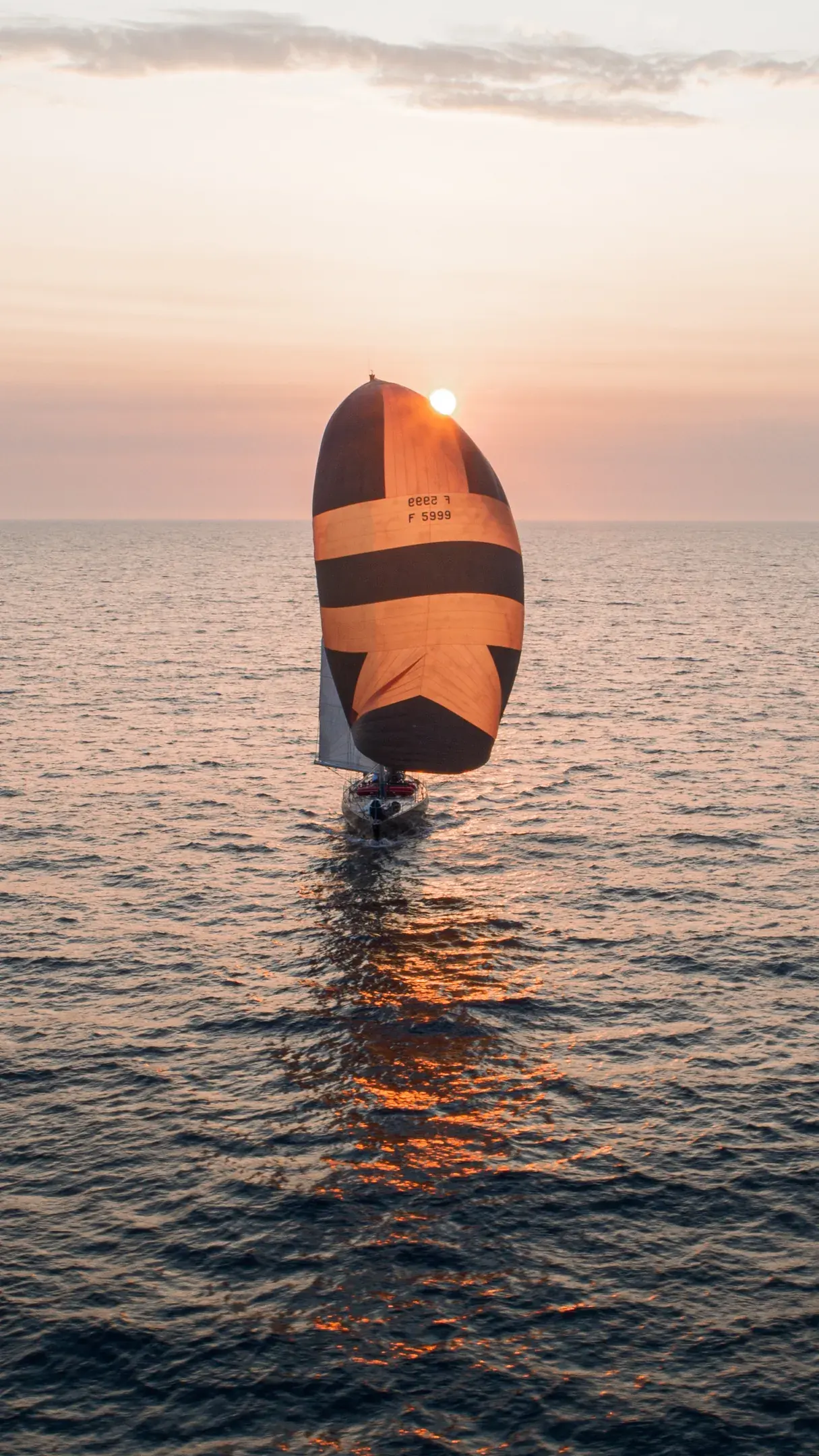 Voilier sous spinnaker orange et noir en pleine mer, face au soleil couchant, vu du ciel